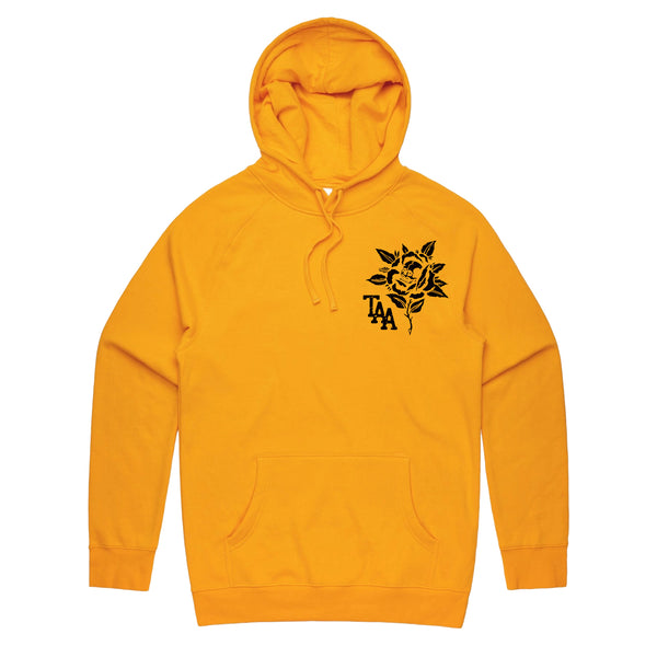 image of the front of a gold pullover hoodie on a white background. the hoodie has a small right chest print in black of a flower with a skull in it and the letters T A A.