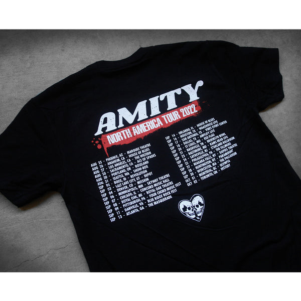 close up angled image of the back of a black tee shirt laid flat on a concrete floor. the tee  has a full back print of the tour dates and locations. aross the top says amity north american tour 2022