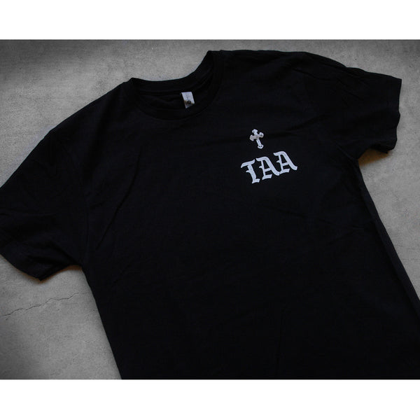 close up angled image of the front of a black tee shirt laid flat on a concrete floor. the tee has a small right chest print in grey that has a small cross and the letters T A A. 