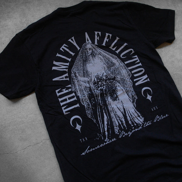 close up, angled image of the back of a black tee shirt laid flat on a concrete floor. the tee has a full back print in grey of a bride with a veil. arched around the bride says the amity affliction. in cursive across the bottom says somewhere beyond the blue