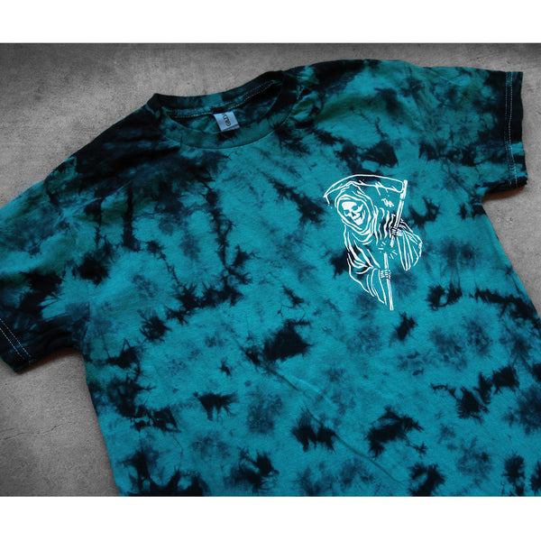 image of a teal and black tie dye tee shirt laid flat on a concrete floor. the front of the tee has a small right chest print in white of a grim reaper