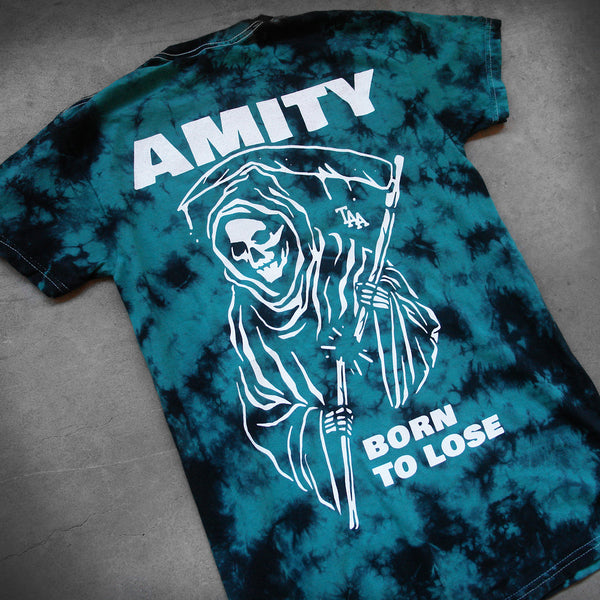 image of the back of a black and teal tie dye tee shirt laid flat on a concrete floor. the back of the tee has a full back print in white of a grim reaper. at the top says amity and the bottom says born to lose
