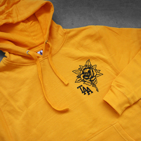 close up angled image of the front of a gold pullover hoodie laid flat on a concrete floor. the hoodie  has a small right chest print in black of a flower with a skull in it and the letters T A A.