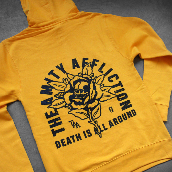 close up angled image of the back of a gold pullover hoodie laid flat on a concrete floor. the hoodie has a full back print in black of a flower with a skull in the center. arched around says the amity affliction. across the bottom says death is all around us